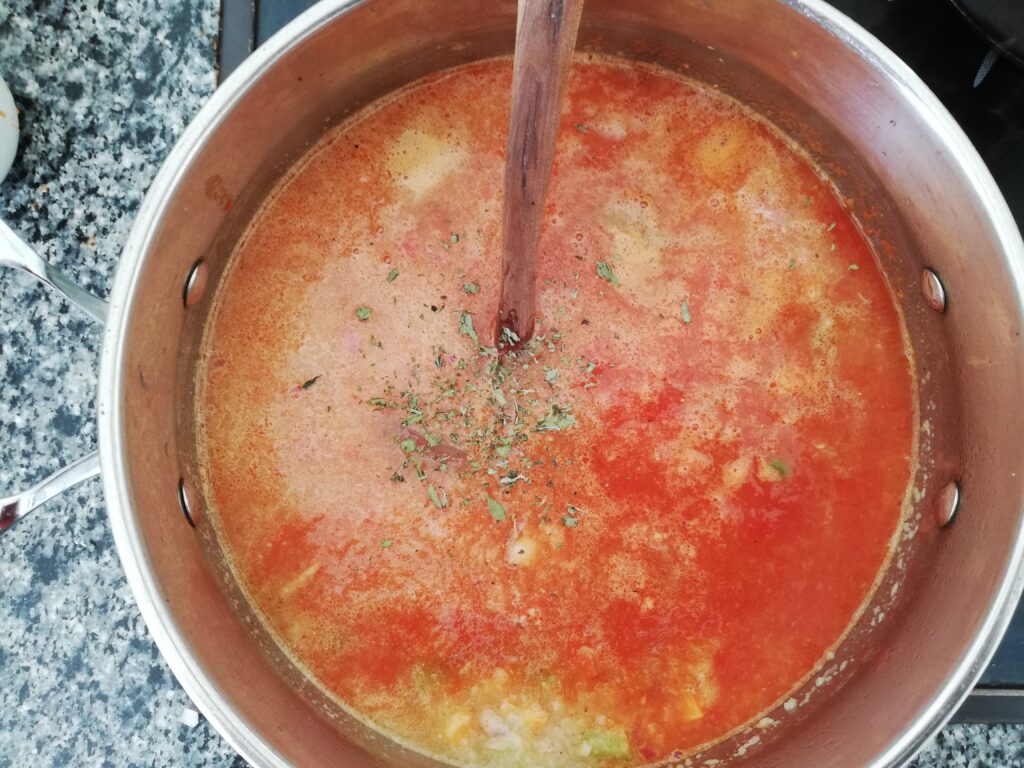 a metal pot with vegetables tomato sauce and stock with dried oregano on top and a wooden spoon