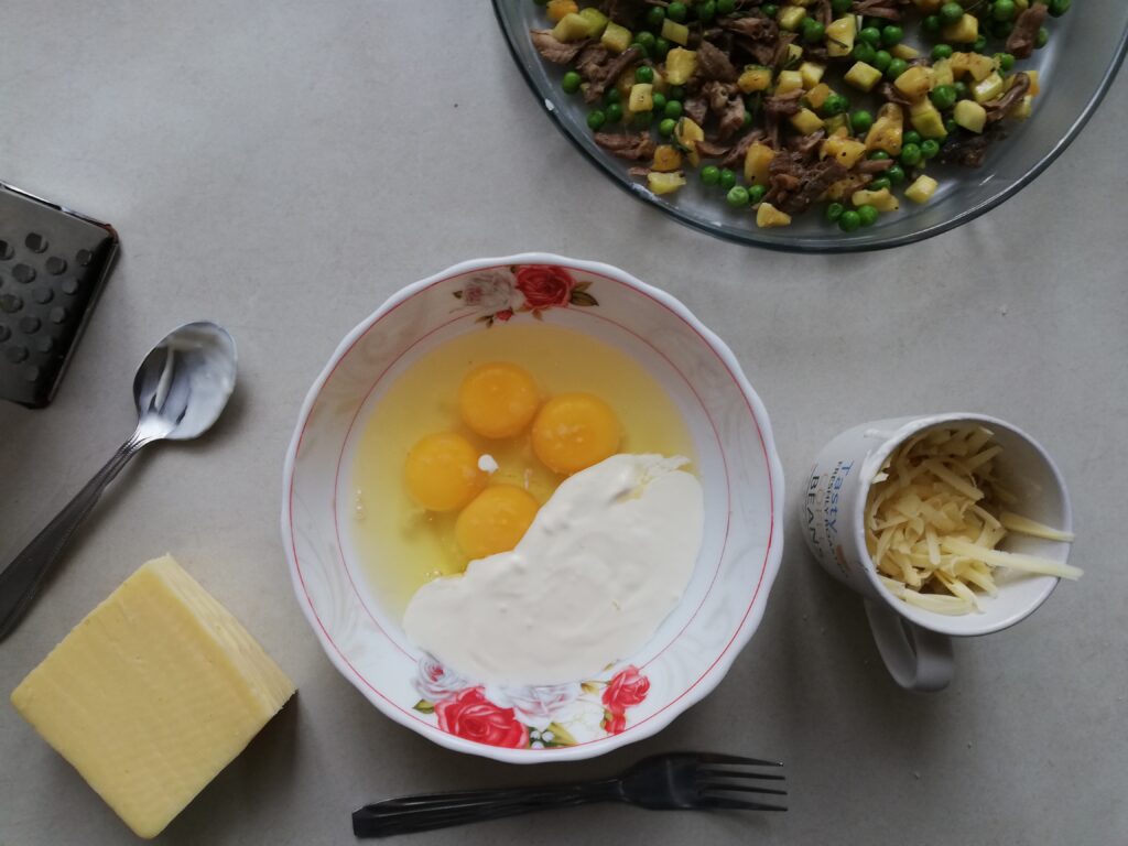 round bowl with cracked eggs and cream block of cheese a grater a round pie dish with cubed courgettes peas and beef a cup half full with shredded cheese a fork and a spoon