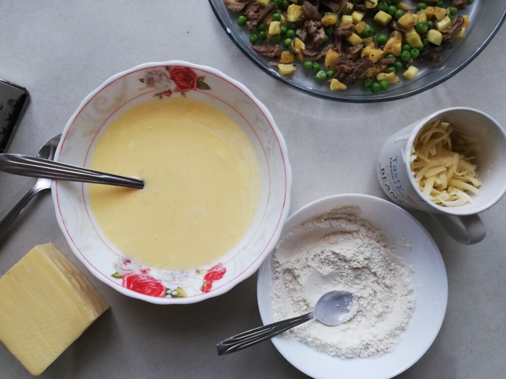 a bowl of whisked eggs and cream a block of cheese a pie dish with cubed courgette peas and beef a cup of shredded cheese and a bowl half full of white flour