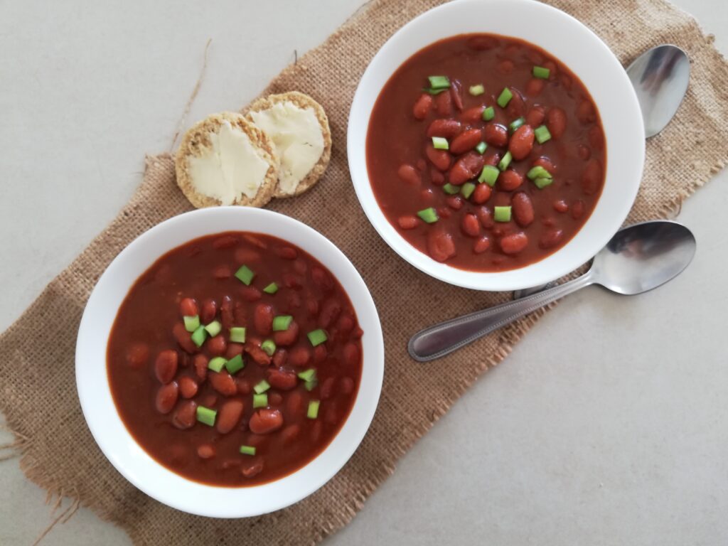 two bowls of baked beans garnished with green onions two metal spoons and a buttered sourdough biscuit on the side