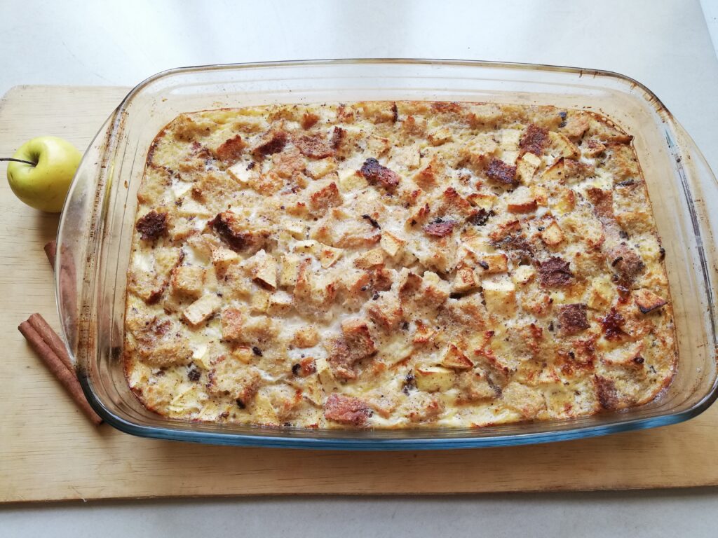 glass casserole dish with sourdough cinnamon and apple baked French toast on a wooden tray with an apple and two cinnamon sticks on the side