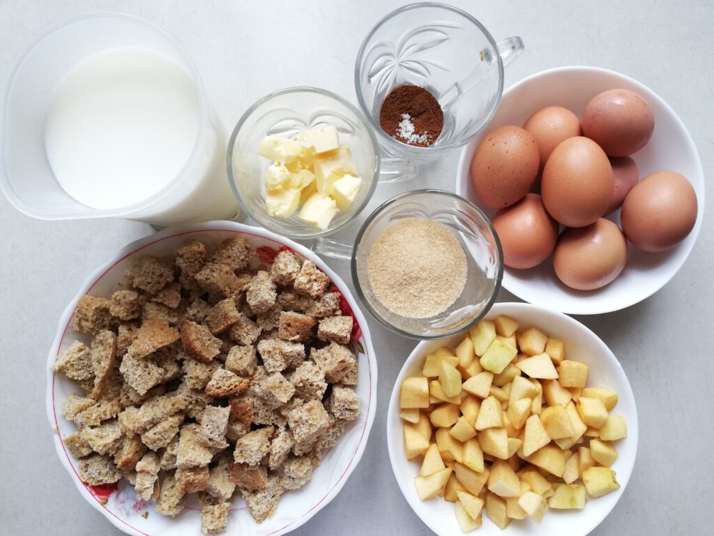 a pitcher of milk a cup of cubed butter a cup with cinnamon and salt a bowl of eggs a cup of brown sugar a bowl of chopped apples a bowl of cubed whole wheat sourdough bread