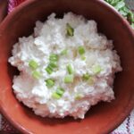 brown bowl with raw milk cottage cheese garnished with green onions with a checkered napkin underneath