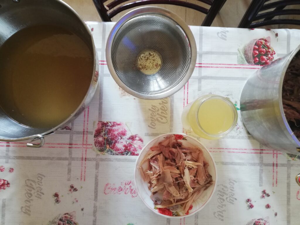 a large pot with chicken broth a funnel a jar of broth and a bowl of shredded chicken meat standing on a kitchen table