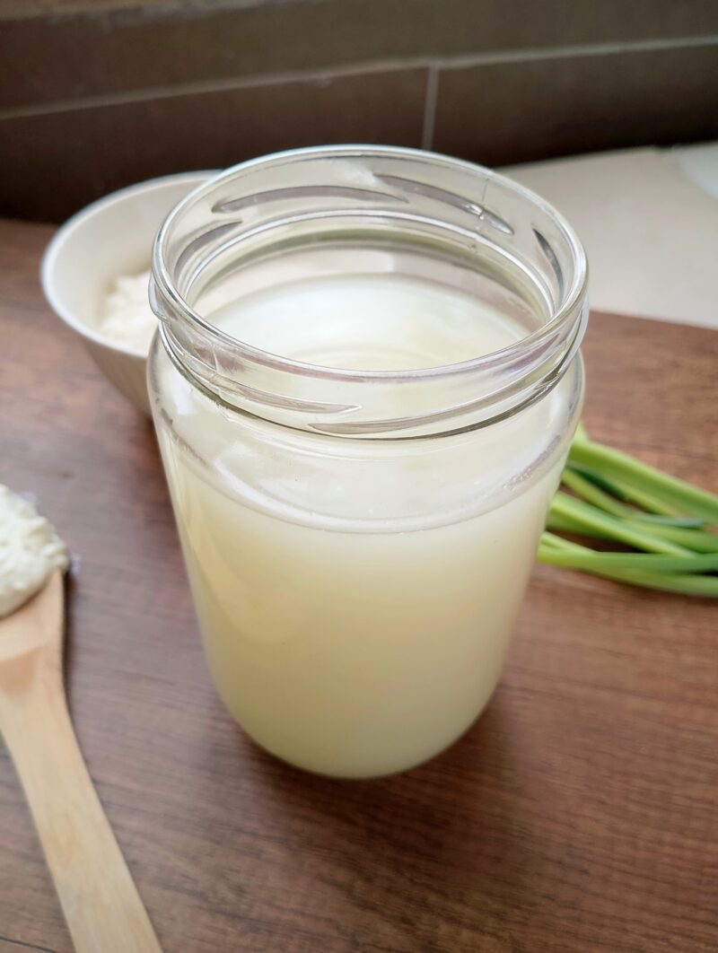 Jar of whey on a wooden board with a spoon of homemade cottage cheese on the side