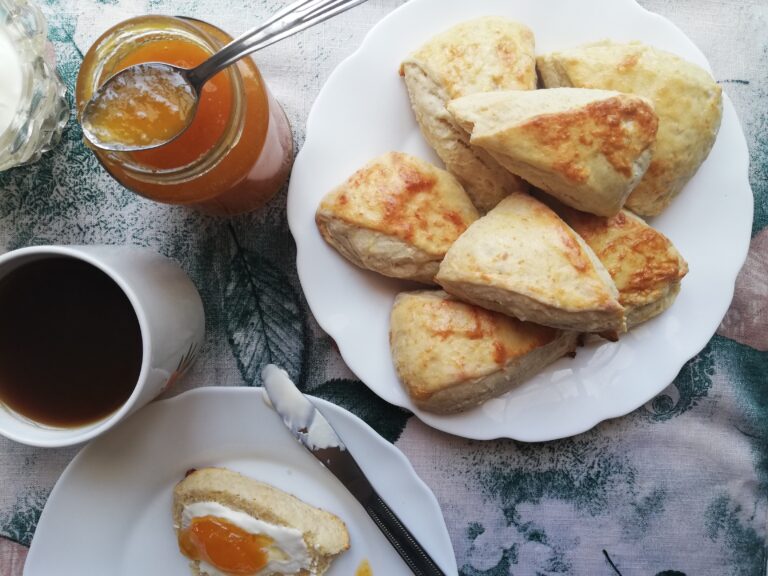 jar of apricot jam a plate of sourdough scones a plate with a butter knife and a scone topped with cream and jam and a cup of tea