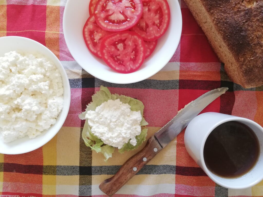 a white bowl of cottage cheese a bowl of sliced tomatoes a loaf of sourdough bread a slice of sourdough bread with lettuce and cottage cheese a buttered bread knife and a mug of tea