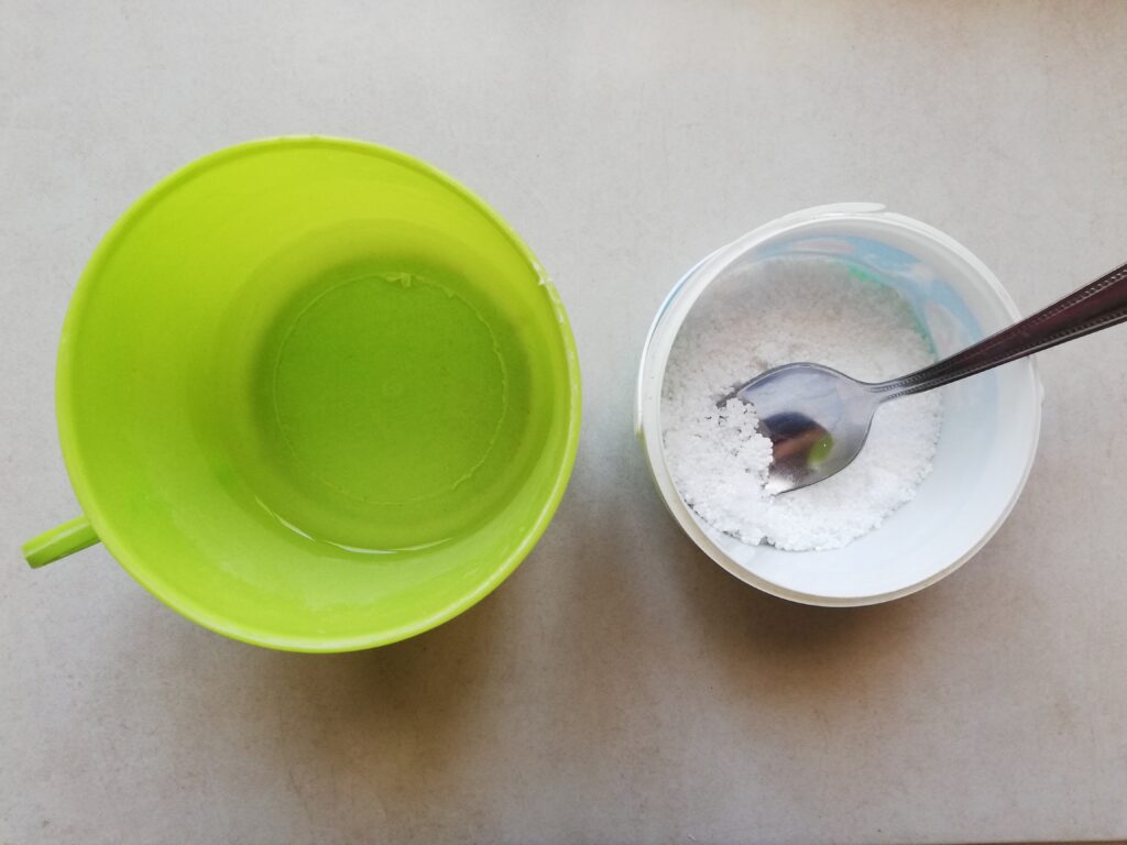green plastic cup with water on the left small plastic contaner with lye crystals and a metal spoon on the right