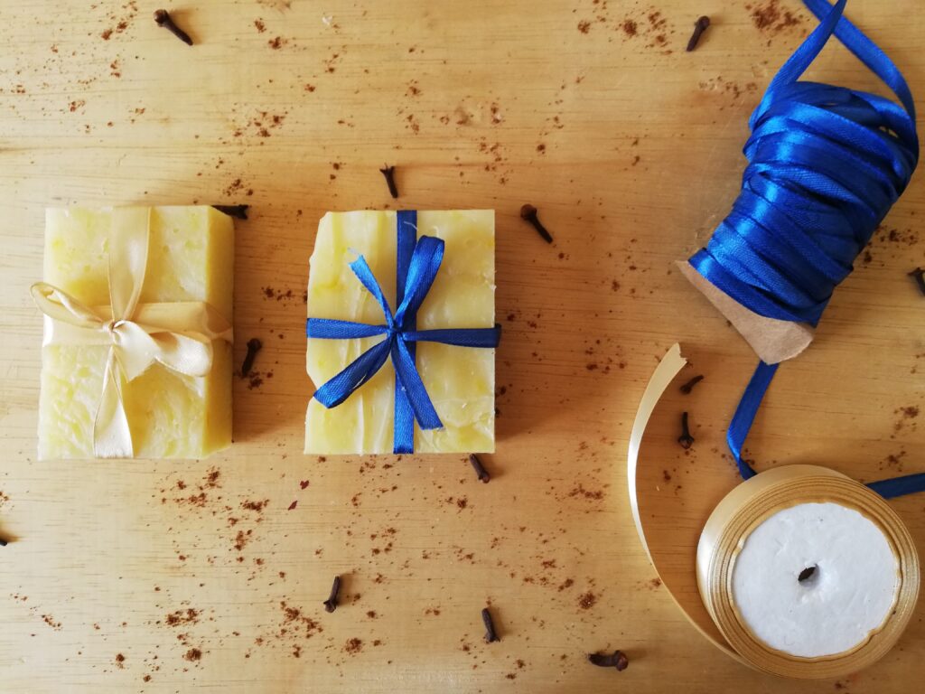 two bars of pumpkin spice soap on a wooden board with cinnamon and cloves sprinkled around on tied with a golden ribbon and one with blue ribbon with a roll of blue ribbon and a roll of golden ribbon on the side