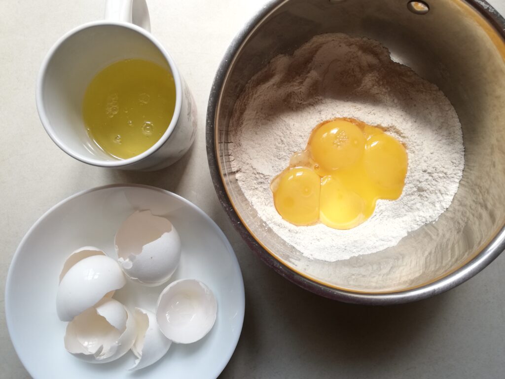 a cup of egg whites a metal mixing bowl with flour and egg yolks and a white bowl with egg shells