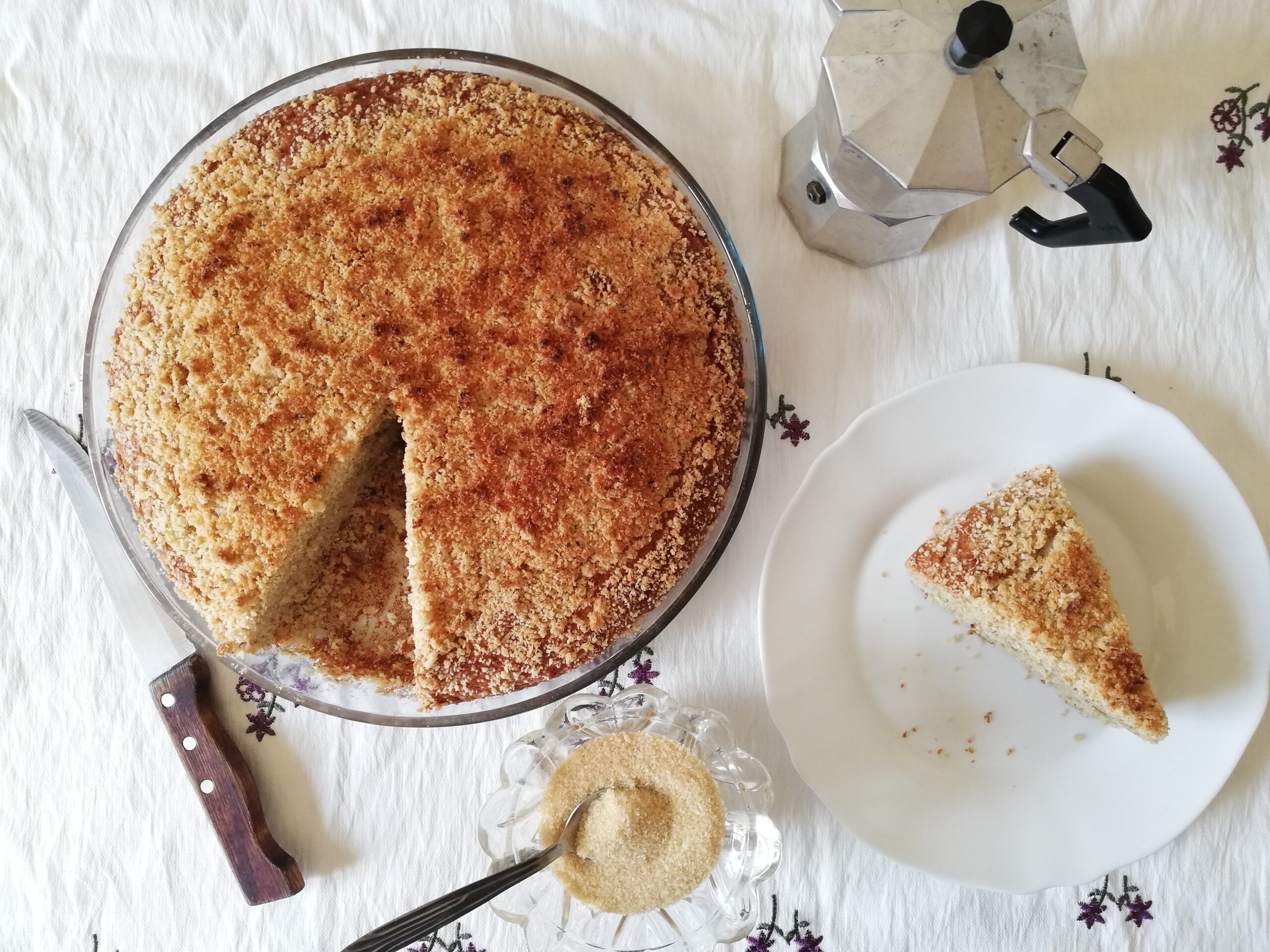 glass round dish with sourdough discard banana and coconut cake with crumb topping a slice of cake on a plate a bowl of brown sugar with a spoon a metal coffee pot and a kitchen knife on the side