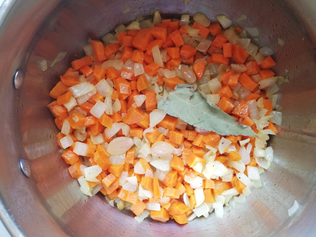 chopped carrots onions garlic cloves and a bay leaf sauteing in a pan