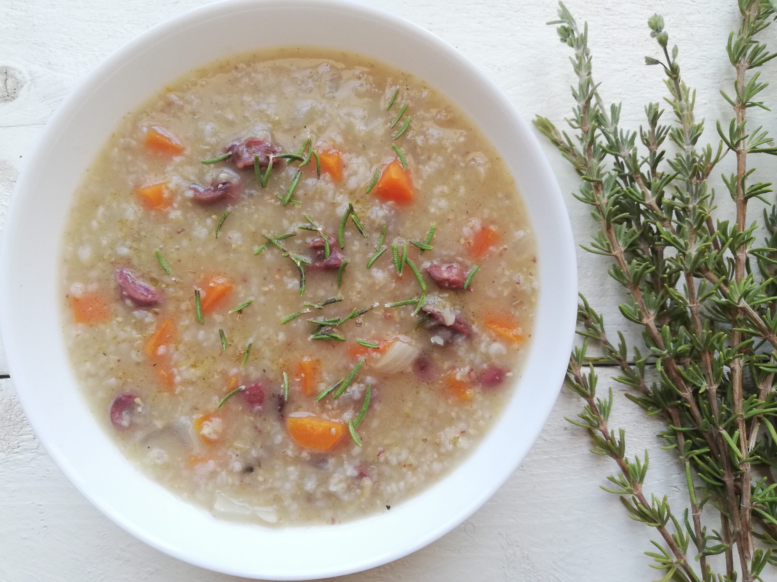 a bowl of barley soup with chicken hearts in a white bowl with some fresh rosemary