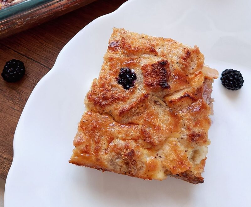 Square of sourdough French toast casserole on plate with some blackberries scattered around