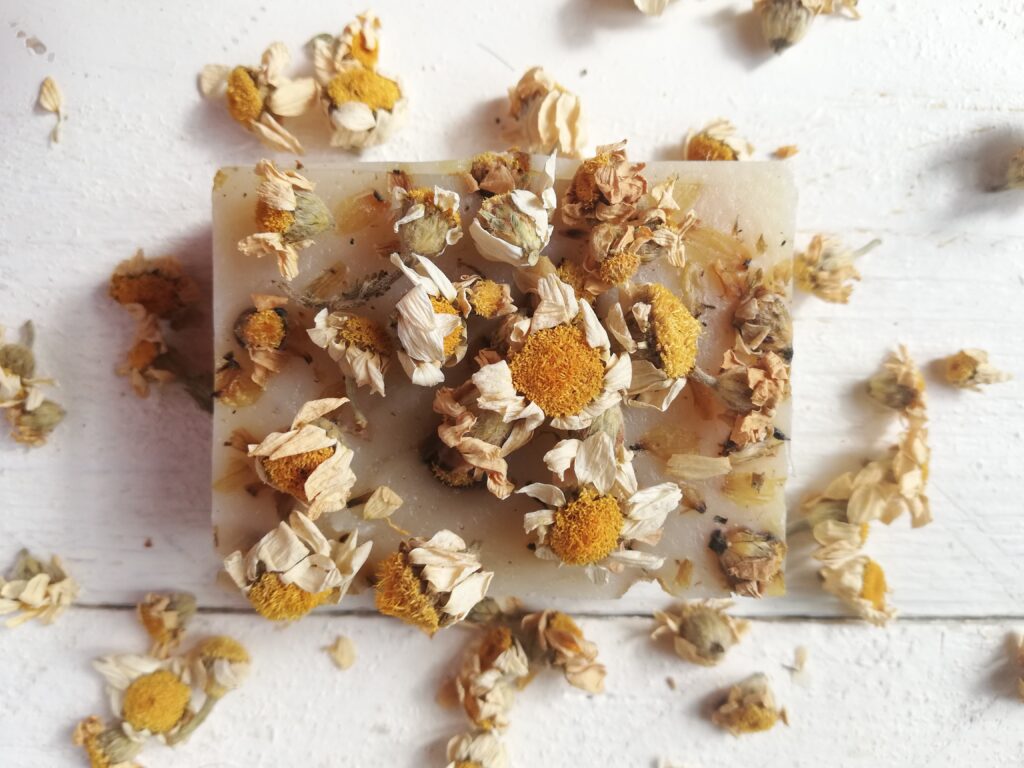 A bar of chamomile cold process soap with chamomile flowers scattered on top and around it