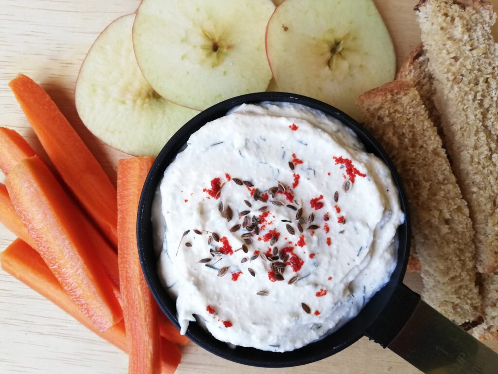 Cottage cheese dip sprinkled with paprika and dill seeds and carrots sticks apple slices and sourdough bread sticks around it