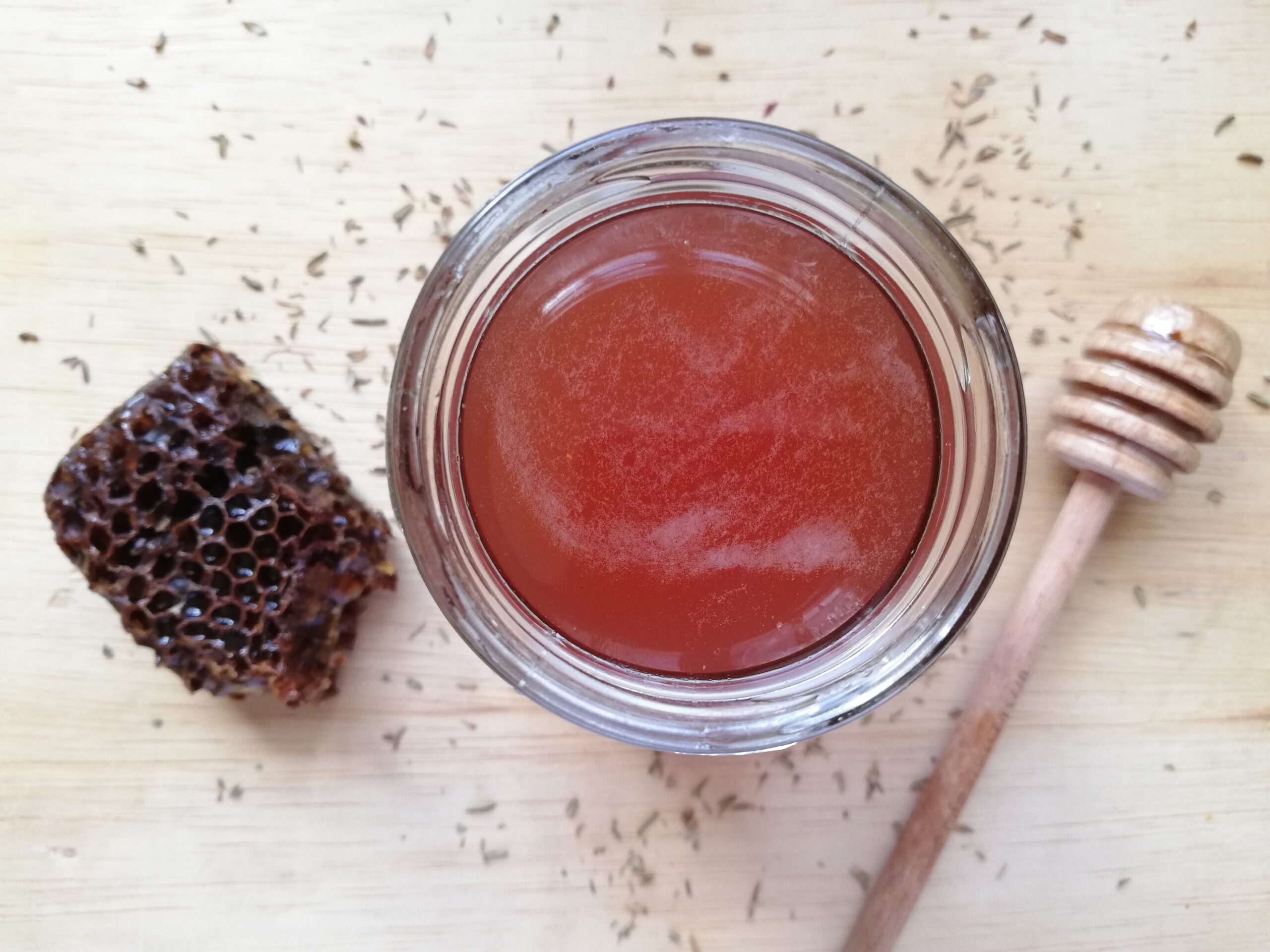 A jar of thyme honey syrup with some thyme sprinkled around a piece of honeycomb and a honey dipper