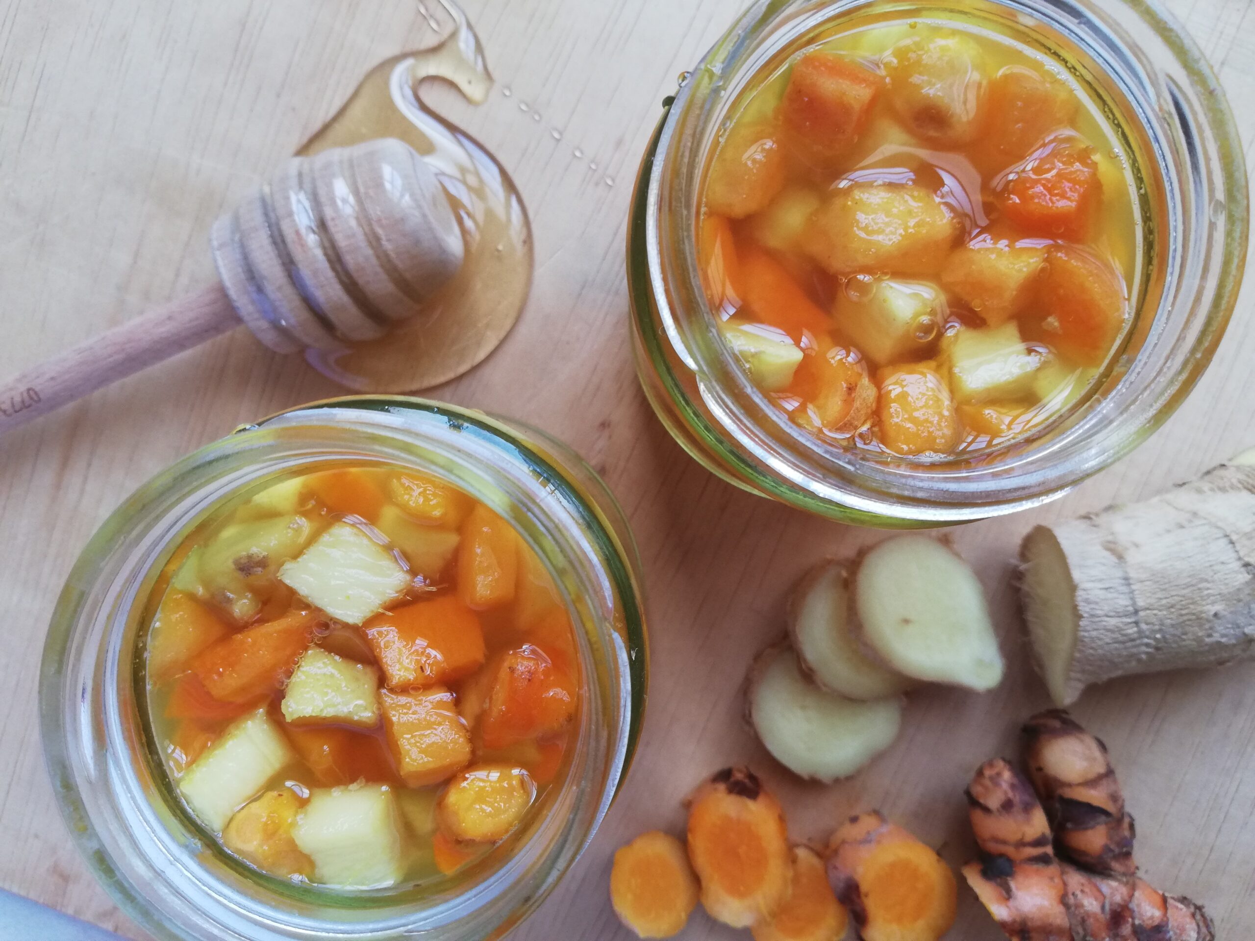 Two jars of fermented turmeric and ginger honey with sliced fresh roots and a honey dipper on a wooden surface