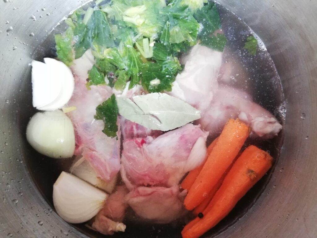 Ingredients for bone broth: chicken bones, onion, carrots, celery, and bay leaf covered with water in a pot