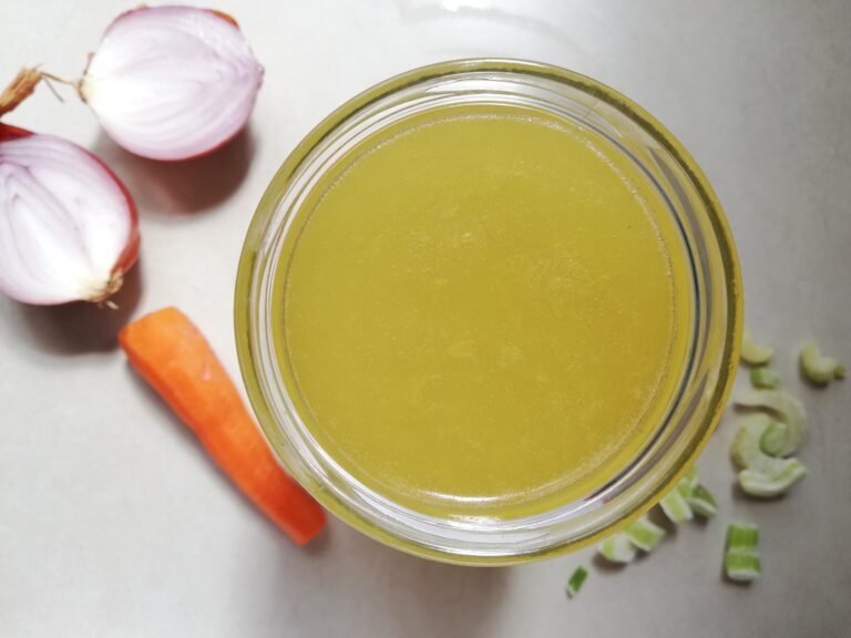 A jar of chicken bone broth on a marble counter top with onion halves, a carrot and some sliced celery around it