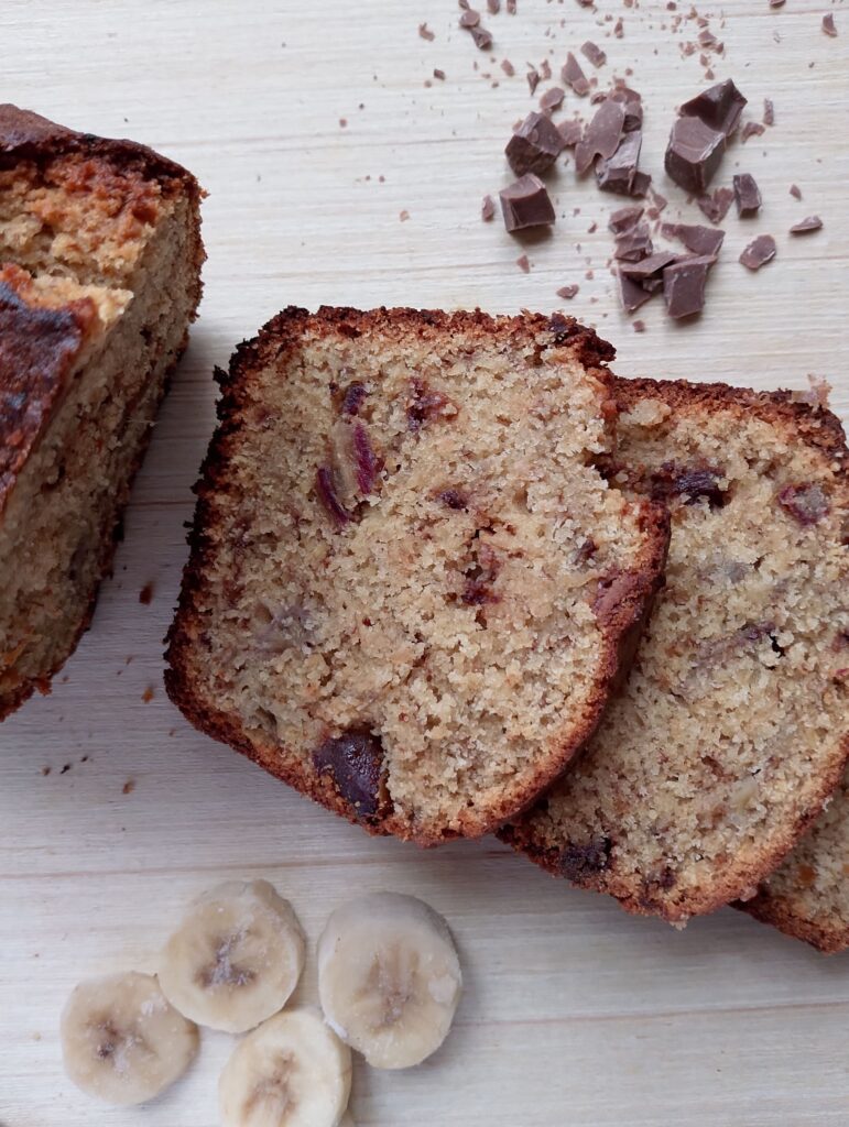 Sourdough discard banana bread sliced on a wooden table with some banana slices and chopped chocolate beside it