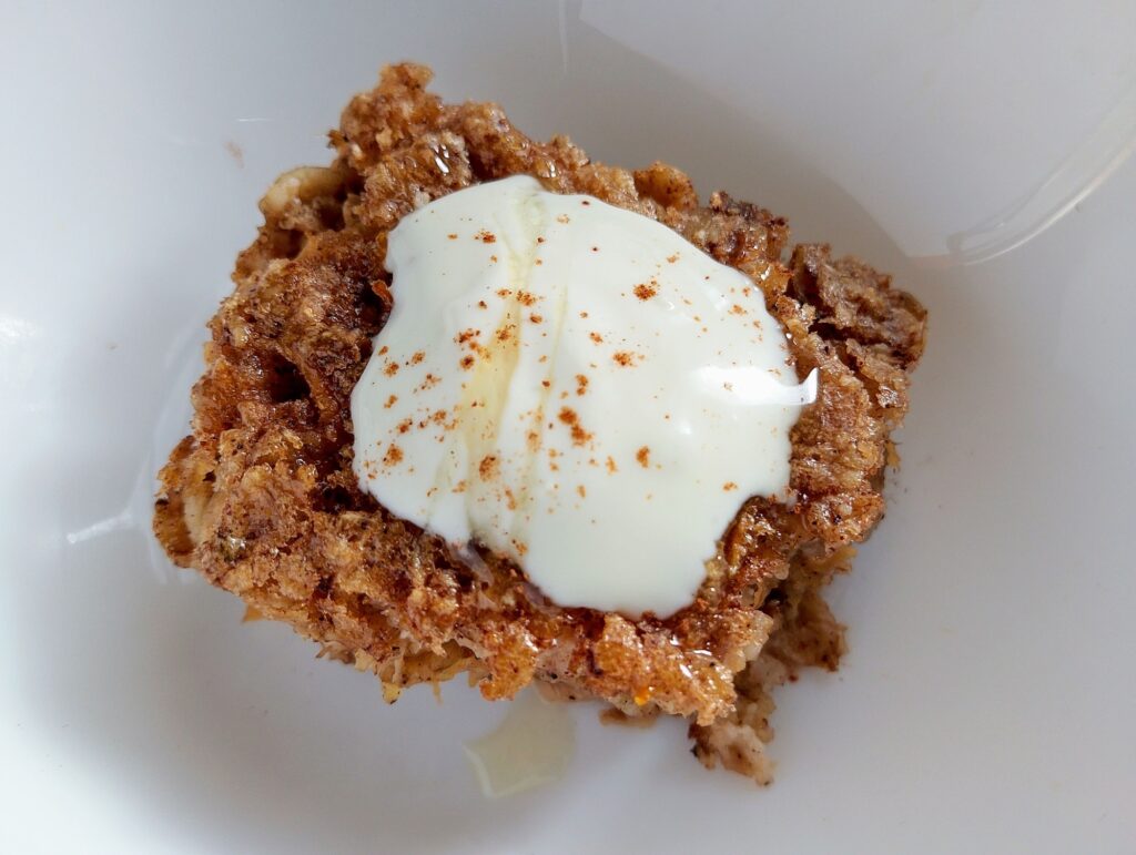 A square slice of carrot cake baked oatmeal with a dollop of cream, a sprinkle of cinnamon, and a drizzle of honey on top