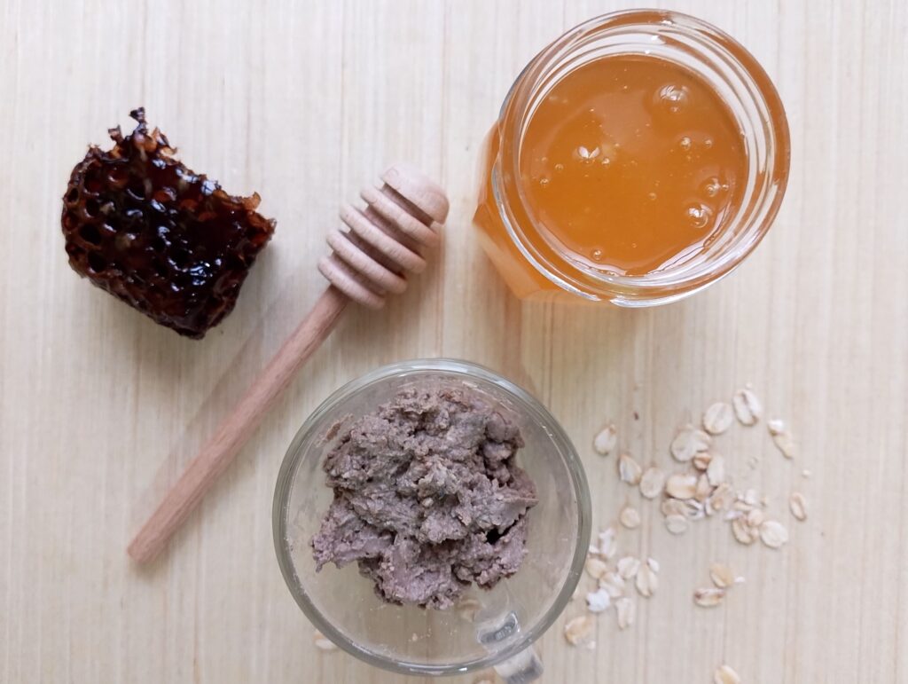 Ready to use DIY clay and oatmeal face mask with honey, with a jar of honey, a honey dipper, some oatmeal, and a piece of honeycomb on the side