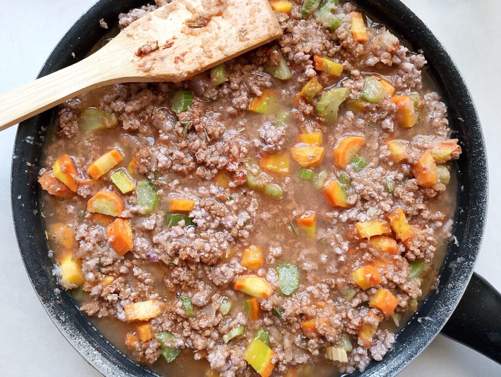 Adding sauteed onion, carrots, and celery as well as the broth to the ground beef in the pan to create the cottage pie filling
