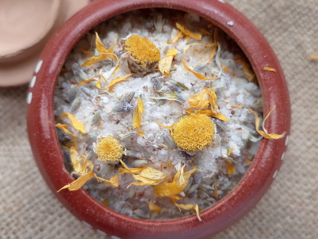 Homemade herbal bath salts in a small clay pot with some calendula, lavender, and chamomile on top