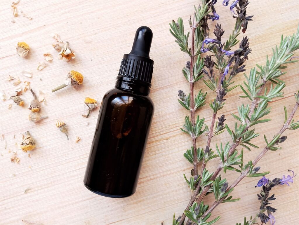 DIY hair and scalp oil in dark amber bottle with some chamomile blossoms and a few sprigs of fresh rosemary on the side