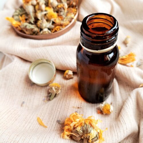 DIY herbal body oil in a dark amber bottle with a small dish of dried calendula on the side