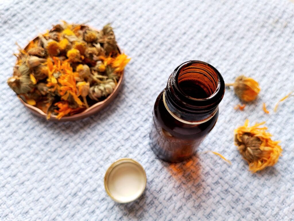 DIY herbal body oil in a dark amber bottle with a small dish of dried calendula blossoms on the side