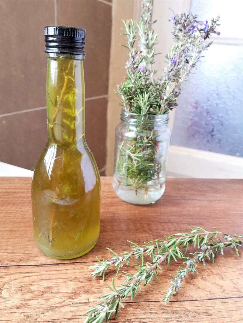 Rosemary infused oil in a bottle with  some fresh rosemary sprigs and blossoms in a jar on the side