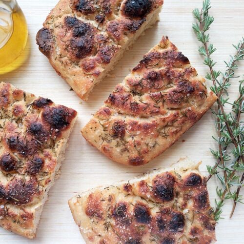 Overnight sourdough focaccia sliced on a wooden table with some fresh rosemary and a jug of olive oil on the side