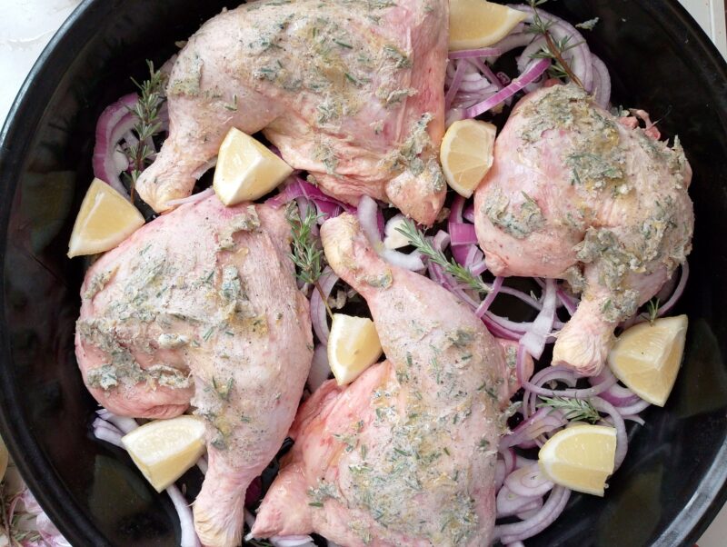 Adding sliced onion, lemon wedges and fresh rosemary sprigs around the marinated chicken legs in the pan