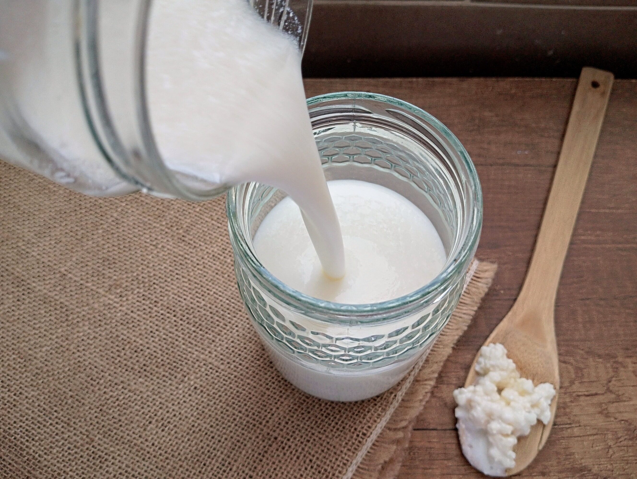 Pouring milk kefir into jar on wooden table, with spoon of kefir grains nearby