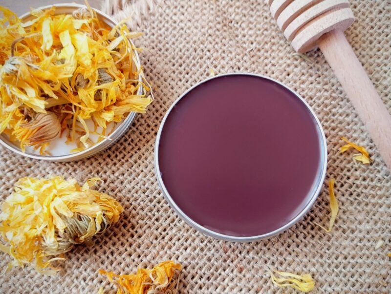 Calendula tallow lip balm in tin on burlap background, with dried calendula petals around