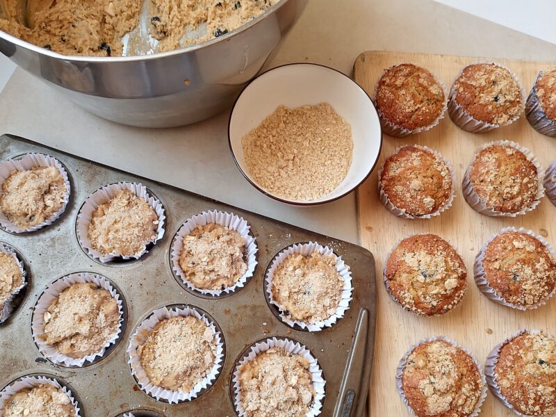 Muffin pan with unbaked muffins, bowl of batter, bowl of streusel topping, and baked muffins cooling on wooden tray