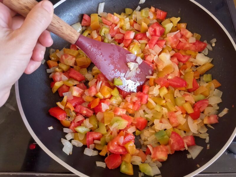 Hand holding spoon, stirring chopped onions, peppers, and tomatoes cooking in the pan