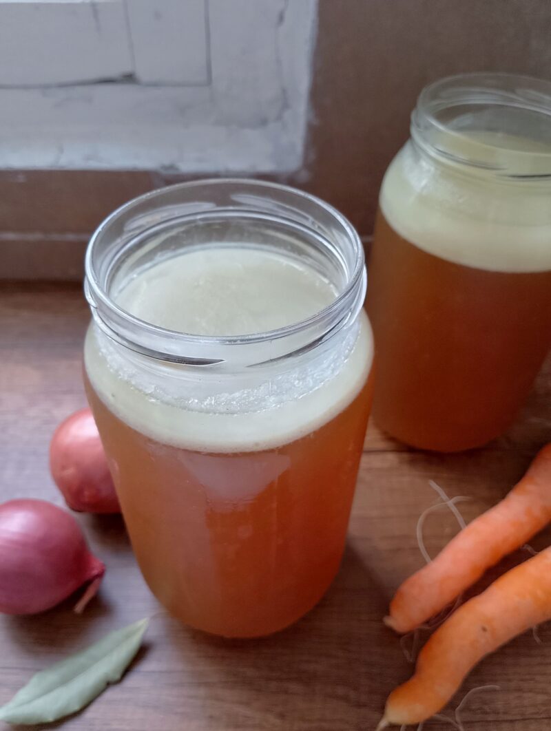 Jars of lamb bone broth with layer of fat on top on wooden table with carrots and onions around